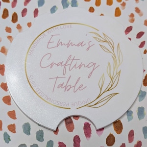 May include: A white, scalloped-edge car coaster with the text "Emma's Crafting Table" in pink script, surrounded by a gold floral design. The words "Handmade with Love" and "Unique Personalised Gifts" are also printed on the coaster.