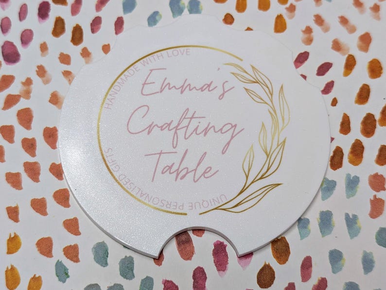 May include: A white, scalloped-edge car coaster with the text "Emma's Crafting Table" in pink script, surrounded by a gold floral design. The words "Handmade with Love" and "Unique Personalised Gifts" are also printed on the coaster.