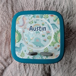 May include: A teal square lunchbox with a colorful dinosaur pattern. The lid features a circular design with the name "Austin" and illustrations of dinosaurs, rainbows, and cupcakes. The lunchbox has two gray latches.