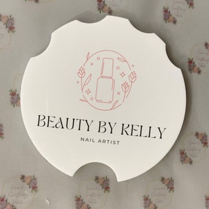 May include: A white, scalloped-edge coaster with the text "BEAUTY BY KELLY NAIL ARTIST" in black. A pink line drawing of a nail polish bottle is in the center, surrounded by floral elements. The background has a repeating floral pattern.