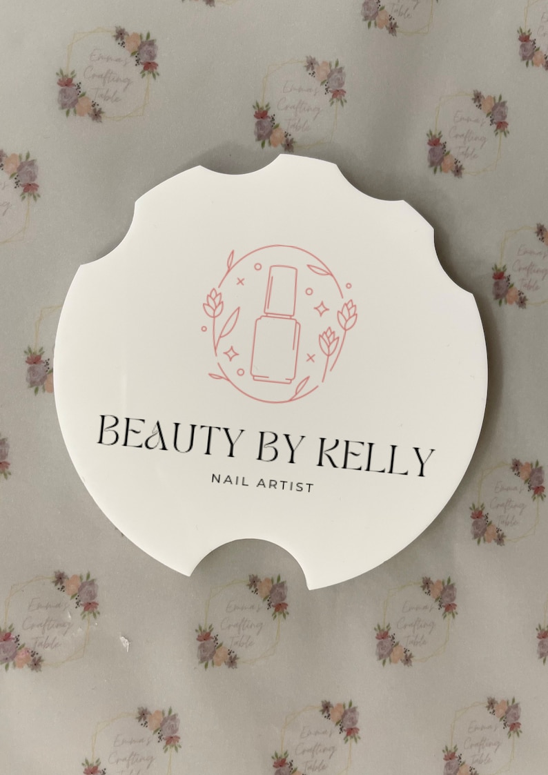 May include: A white, scalloped-edge coaster with the text "BEAUTY BY KELLY NAIL ARTIST" in black. A pink line drawing of a nail polish bottle is in the center, surrounded by floral elements. The background has a repeating floral pattern.