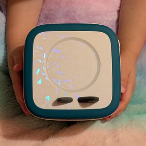 May include: A square, teal and white device with a circular center and two black buttons. The center has a shimmering, iridescent design. The device is held by two hands against a fluffy, multicolored background.