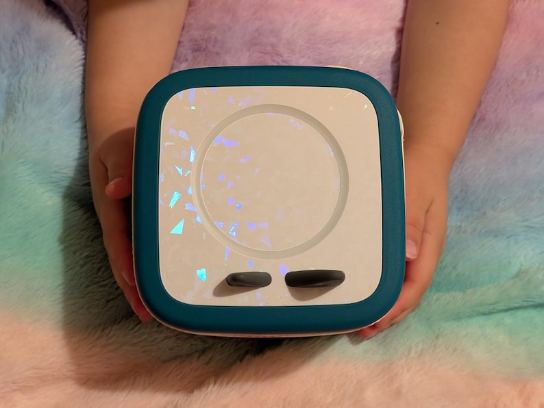 May include: A square, teal and white device with a circular center and two black buttons. The center has a shimmering, iridescent design. The device is held by two hands against a fluffy, multicolored background.