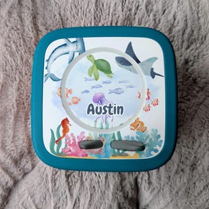 May include: A teal square container with a white lid featuring an underwater scene. The lid has illustrations of a shark, turtle, fish, and a jellyfish. The name "Austin" is written in the centre of the lid.