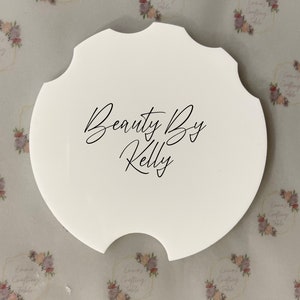 May include: A white, scalloped-edge coaster with the text "Beauty By Kelly" in black script. The coaster has a unique shape and is likely designed to fit in a car's cup holder. The background features a floral pattern.