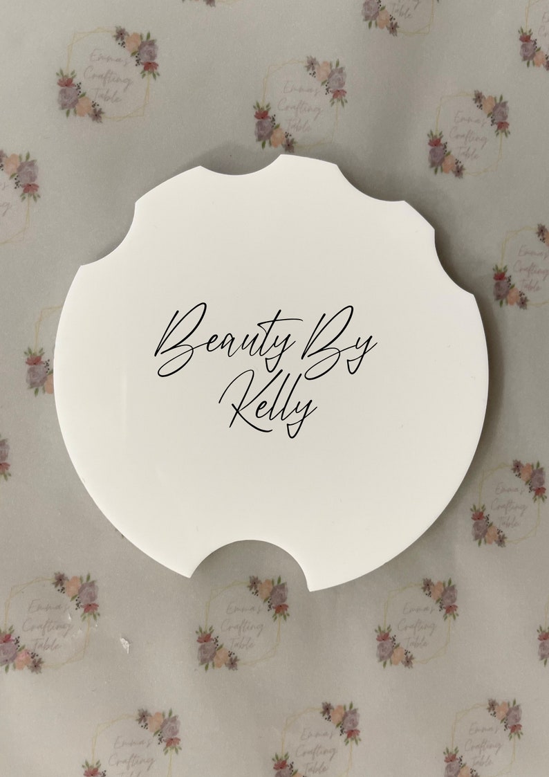 May include: A white, scalloped-edge coaster with the text "Beauty By Kelly" in black script. The coaster has a unique shape and is likely designed to fit in a car's cup holder. The background features a floral pattern.
