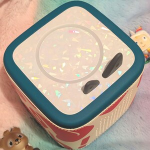 May include: A square, white and teal audio player with a holographic design on top. The sides are patterned with red hearts and white accents. Two black buttons are visible on the top. A small toy is visible in the background.