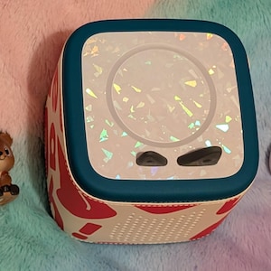 May include: A white and red patterned speaker with a teal border, featuring a holographic design on the front. Two small black buttons are visible below the screen. Two small figurines, a deer and a unicorn, are on either side of the speaker.