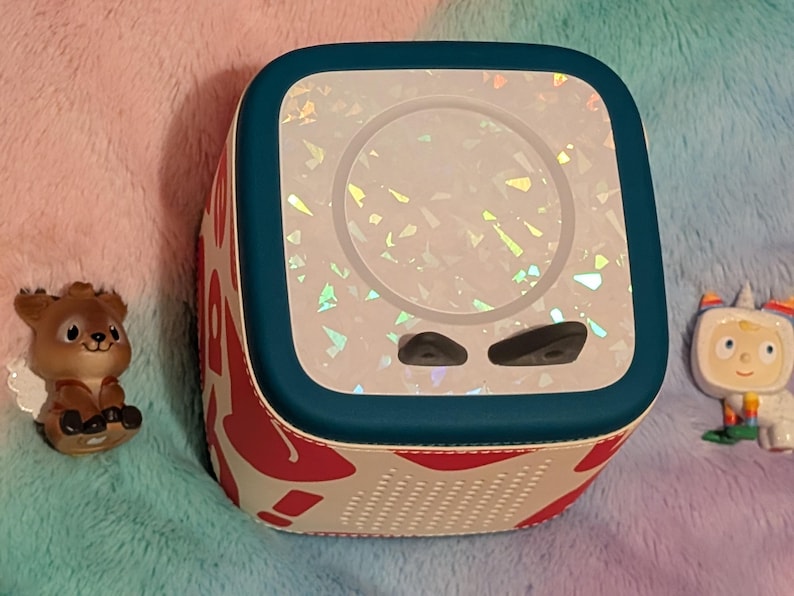 May include: A white and red patterned speaker with a teal border, featuring a holographic design on the front. Two small black buttons are visible below the screen. Two small figurines, a deer and a unicorn, are on either side of the speaker.