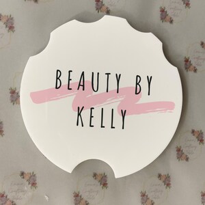 May include: White car coaster with a unique scalloped edge. The coaster features the text "BEAUTY BY KELLY" in black, with a pink brushstroke accent. A decorative item for a car.