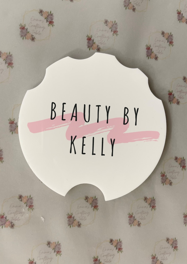 May include: White car coaster with a unique scalloped edge. The coaster features the text "BEAUTY BY KELLY" in black, with a pink brushstroke accent. A decorative item for a car.