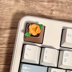 May include: Close-up of a white mechanical keyboard with light blue and white keycaps. The escape key is replaced with a custom keycap featuring a brown base and an orange flower design. The keyboard is on a wooden surface.