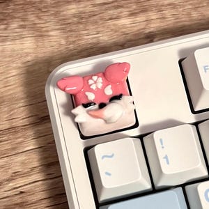 May include: A white mechanical keyboard with light blue and white keycaps. A custom keycap featuring a pink fox design with white accents is installed on the keyboard. The keyboard has function keys and alphanumeric keys.