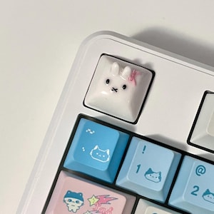 May include: A white mechanical keyboard with custom keycaps. The keycaps feature various designs, including a bunny, cat faces, and the word "Hello!". The color scheme includes white, blue, and pink.