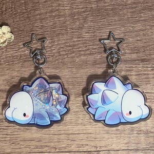 May include: Two clear acrylic keyrings with a cartoon hedgehog design in white, blue, and purple. Each keyring has a silver star-shaped clasp. The keyrings are displayed on a wooden surface with decorative flowers.