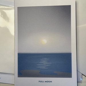 May include: A greeting card with a minimalist illustration of a full moon over the ocean. The card features a gradient sky, a yellow moon, and dark blue water. The words "FULL MOON" are printed at the bottom. A pen rests in front of the card.