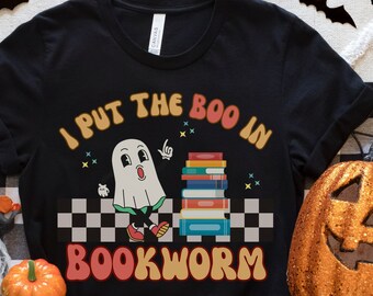 May include: Black t-shirt featuring a white ghost holding a stack of books. The text on the shirt reads "I put the boo in bookworm".