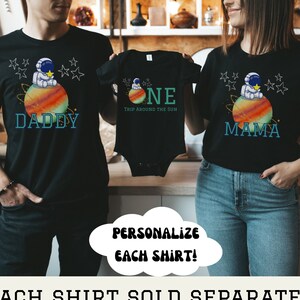 Puede incluir: Un conjunto de tres camisetas negras con un diseño de temática espacial. Las camisetas presentan un astronauta sentado en un planeta con estrellas y el texto "Papá", "Un viaje alrededor del sol", y "Mamá".