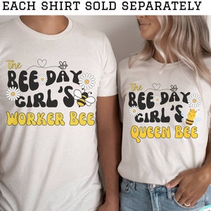 May include: Two white t-shirts with bee-themed designs. The first shirt says "The Bee Day Girl's Worker Bee" with a bee and daisies. The second shirt says "The Bee Day Girl's Queen Bee" with a bee and daisies.