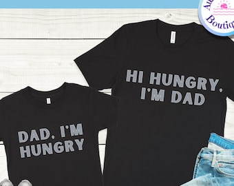 May include: Two black t-shirts with a gray graphic print. The larger shirt says "Hi Hungry, I'm Dad." The smaller shirt says "Dad, I'm Hungry."
