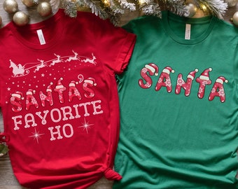 Può includere: Due magliette a tema natalizio. Una maglietta rossa con il testo "Santa's Favorite Ho" in lettere bianche con un cappello di Babbo Natale rosso sulla lettera "A". L'altra maglietta è verde con la parola "SANTA" in lettere bianche con cappelli di Babbo Natale rossi su ogni lettera. Il testo "EACH SHIRT SOLD SEPARATELY." è in basso nell'immagine.