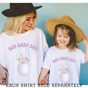 May include: Two white t-shirts with the text "Bad Hare Day" in purple letters and a cartoon bunny egg design. The shirts are worn by a woman and a young girl.