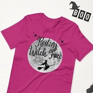 May include: A pink t-shirt featuring the text "Resting Witch Face" and a black silhouette of a witch flying on a broomstick in front of a full moon.