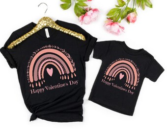 Könnte beinhalten: Zwei schwarze T-Shirts mit einem rosafarbenen Regenbogen-Design und dem Text "Happy Valentine's Day".
