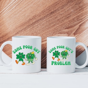 May include: Two white ceramic mugs with a green shamrock and cupcake design. The text on the mugs reads "Some Poor Guy's Problem".