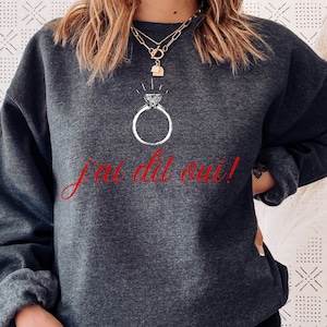 Könnte beinhalten: Ein dunkelgrauer Pullover mit einem weißen Ring- und Diamant-Design und dem Text "j'ai dit oui!" in Rot.