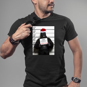 Peut inclure: Un t-shirt noir avec une image blanche et noire d'un gorille portant un chapeau de Père Noël et tenant une pancarte qui dit "ARRESTED FOR DISORDERLY CONDUCT: Got Lost in a Jungle".