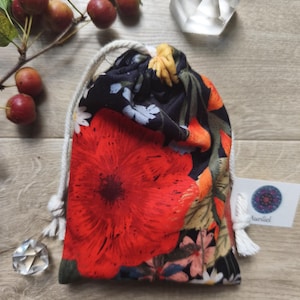 May include: A drawstring pouch with a vibrant floral pattern featuring a large red poppy. The bag has a white drawstring and a small tag with the word "Aunliel". The background includes small red apples and a clear crystal.