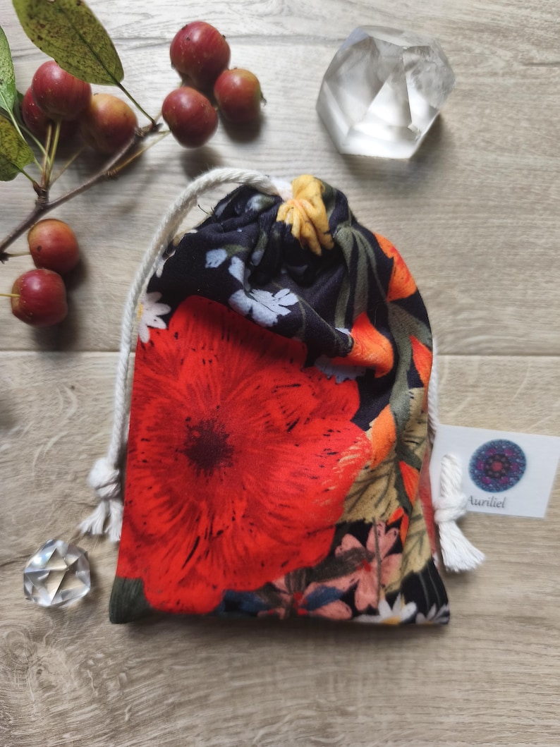 May include: A drawstring pouch with a vibrant floral pattern featuring a large red poppy. The bag has a white drawstring and a small tag with the word "Aunliel". The background includes small red apples and a clear crystal.