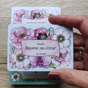 May include: A white card with pink and white flowers and a scroll with the text "Oracle Baume au coeur Par Aurihel".
