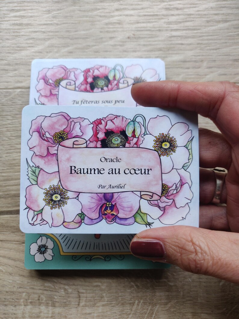 May include: A white card with pink and white flowers and a scroll with the text "Oracle Baume au coeur Par Aurihel".