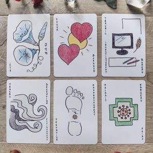 May include: Six illustrated tarot cards with hand-drawn images and French text. The cards feature images of flowers, hearts, a foot, and a computer. The cards are arranged on a wooden surface with crystals and flowers.
