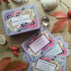 May include: A set of oracle cards with floral illustrations and text in French. The cards are on a wooden surface with a seashell, crystals, and rose hips.
