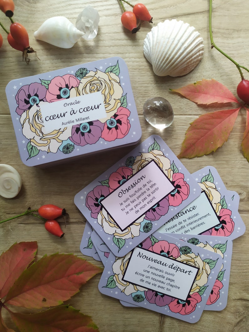 May include: A set of oracle cards with floral illustrations and text in French. The cards are on a wooden surface with a seashell, crystals, and rose hips.