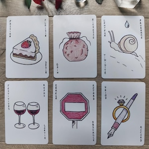 May include: Six tarot cards with hand-drawn illustrations and French text. The cards depict a slice of cake, a money bag, a snail, two wine glasses, a stop sign, and a pen with a ring. The cards are arranged on a wooden surface with decorative elements.