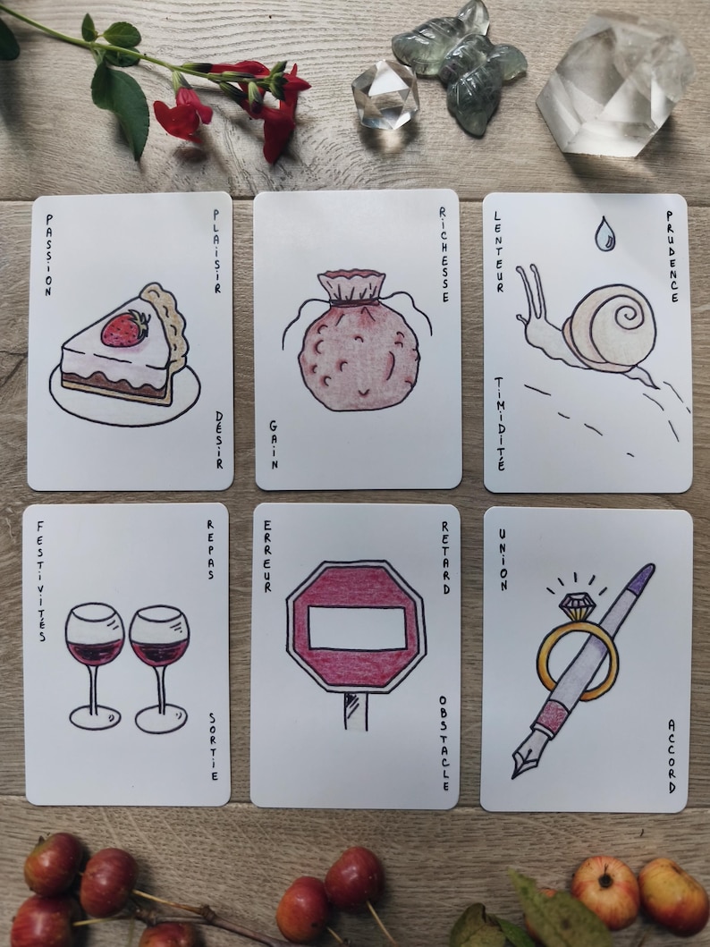 May include: Six tarot cards with hand-drawn illustrations and French text. The cards depict a slice of cake, a money bag, a snail, two wine glasses, a stop sign, and a pen with a ring. The cards are arranged on a wooden surface with decorative elements.