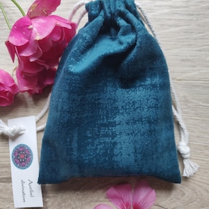 May include: A teal velvet drawstring bag with a white cord. The bag is sitting on a wooden surface with pink flowers on either side. A white tag with the text "Auniel divination" is visible in the lower left corner.