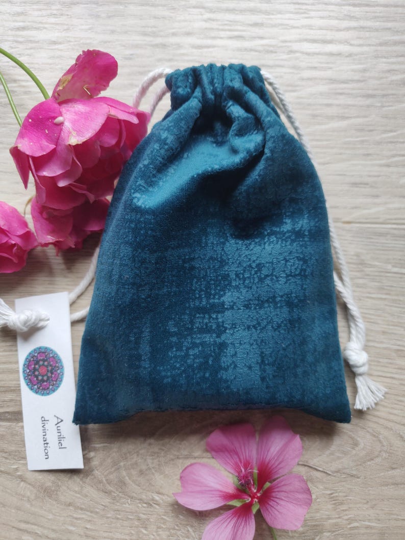 May include: A teal velvet drawstring bag with a white cord. The bag is sitting on a wooden surface with pink flowers on either side. A white tag with the text "Auniel divination" is visible in the lower left corner.