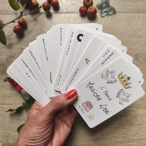 May include: A deck of white oracle cards fanned out in a hand, featuring illustrations of doves, a crown, and a bag. The cards have French text, including "Magicae Libri" and other words like "Maladie" and "Printemps."