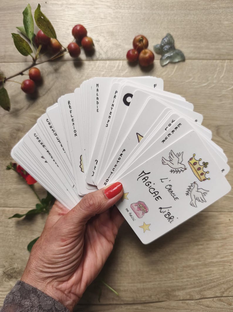 May include: A deck of white oracle cards fanned out in a hand, featuring illustrations of doves, a crown, and a bag. The cards have French text, including "Magicae Libri" and other words like "Maladie" and "Printemps."