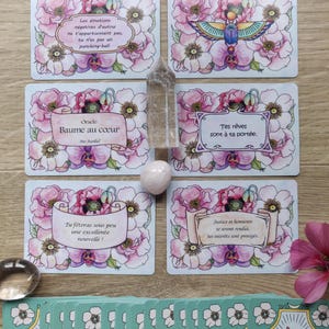 May include: A set of oracle cards with pink floral designs and gold accents. The cards feature inspirational messages in French, such as "Baume au coeur" and "Tes r&ecirc;ves sont &agrave; ta port&eacute;e."