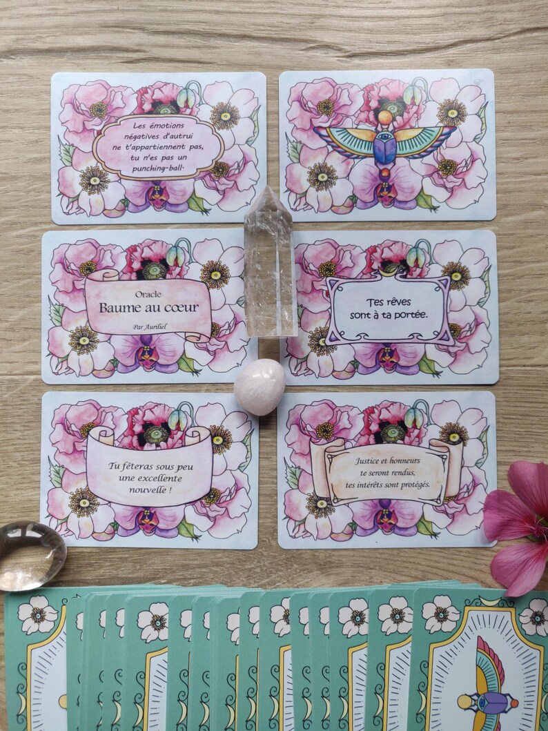 May include: A set of oracle cards with pink floral designs and gold accents. The cards feature inspirational messages in French, such as "Baume au coeur" and "Tes r&ecirc;ves sont &agrave; ta port&eacute;e."