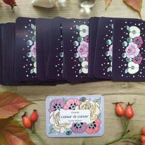 May include: A deck of oracle cards with a purple background and floral illustrations. The cards are stacked on a wooden surface. The box for the cards is in the foreground and features a floral design with the text "Oracle coeur &agrave; coeur Aurilie Millaret".