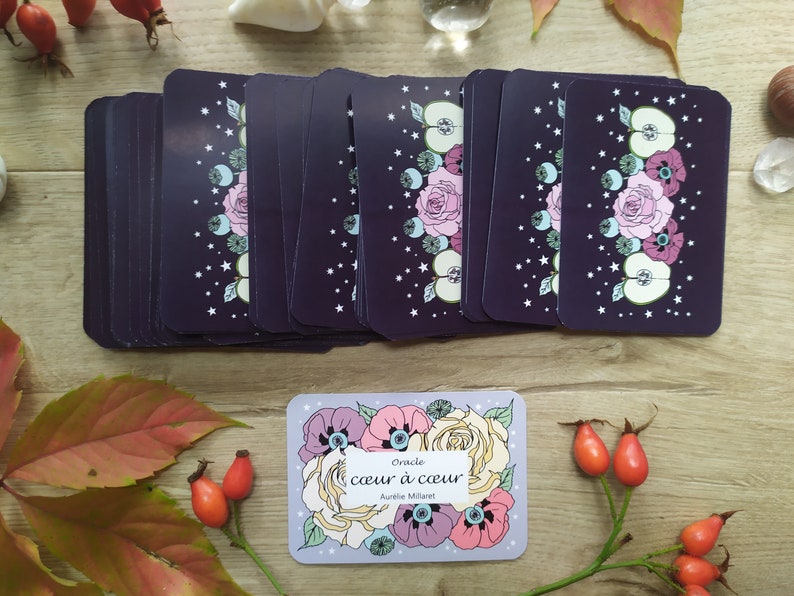 May include: A deck of oracle cards with a purple background and floral illustrations. The cards are stacked on a wooden surface. The box for the cards is in the foreground and features a floral design with the text "Oracle coeur &agrave; coeur Aurilie Millaret".