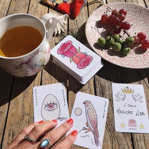 May include: A stack of tarot cards with rose illustrations, a cup of tea, and a plate of grapes on a wooden table. The cards feature various drawings and French text. A small bag with a floral pattern is also visible.