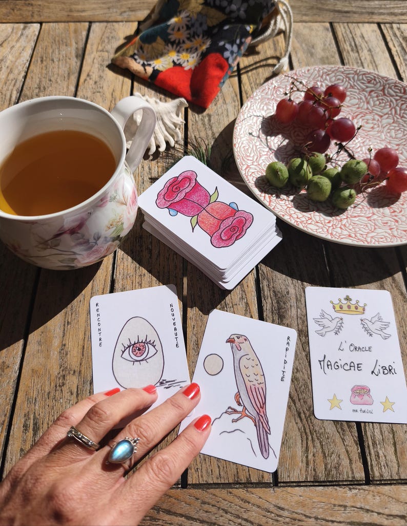 May include: A stack of tarot cards with rose illustrations, a cup of tea, and a plate of grapes on a wooden table. The cards feature various drawings and French text. A small bag with a floral pattern is also visible.